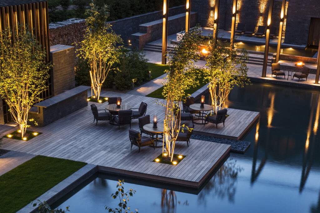 Außenterrasse mit Sitzgelegenheiten, beleuchteten Bäumen und moderner Beleuchtung neben einem spiegelnden Pool in der Abenddämmerung.