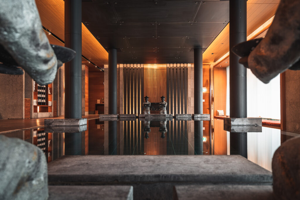 Modernes, ruhiges Interieur mit Statuen, Säulen und spiegelndem Wasser, das zu zwei sitzenden Figuren im Hintergrund führt.