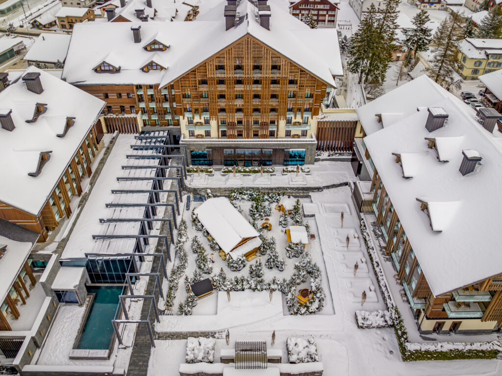 Luftaufnahme einer schneebedeckten Hotelanlage mit Innenhöfen, Holzgebäuden und einem Außenpool.