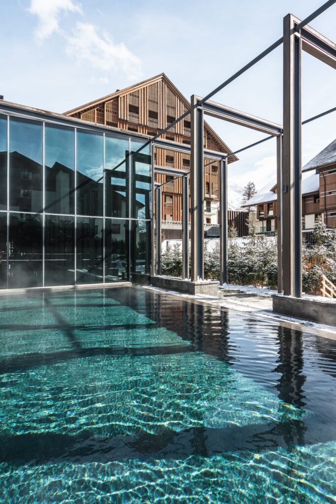 Modernes Glasgebäude mit Außenpool, Holzstrukturen und Berghütten im Hintergrund an einem sonnigen Tag.