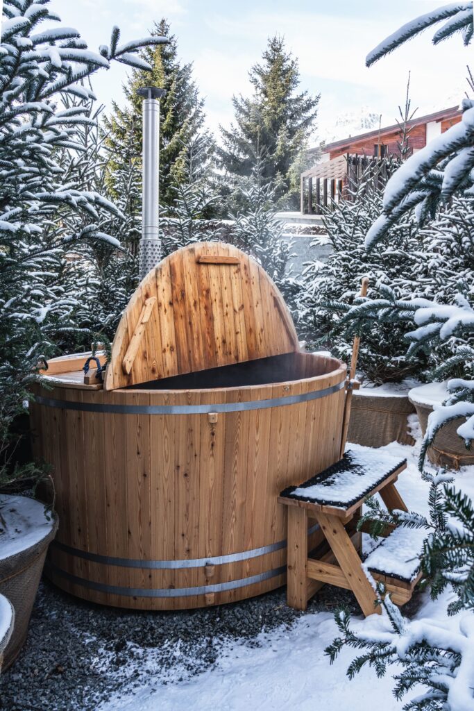 Zwischen schneebedeckten Kiefern und einem kleinen hölzernen Trittschemel steht ein hölzerner Whirlpool mit hochgezogenem Deckel.